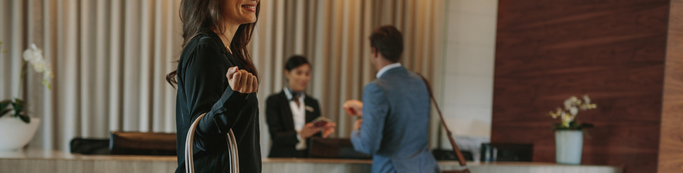 a man checking in to a hotel, talking to a receptionist. A woman with a handbag in the foreground.